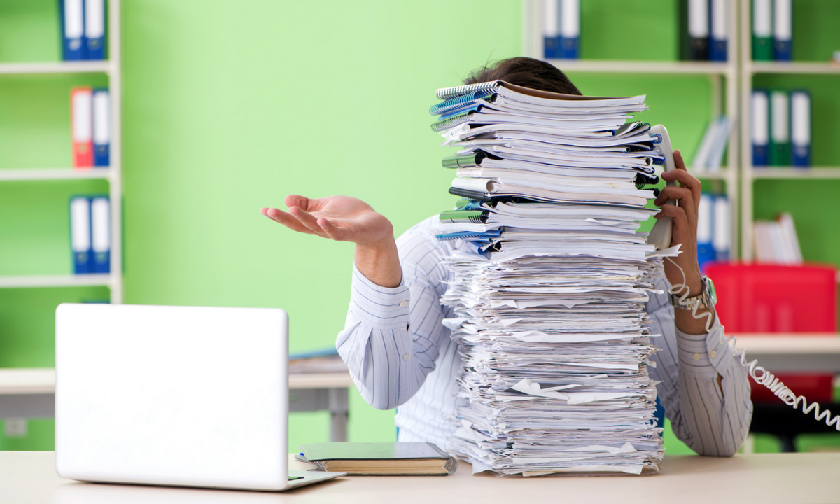 A person in a striped shirt sits at a desk covered by a massive stack of papers, holding a phone receiver to their ear while gesturing with one hand. A laptop is on the desk in front of them, with a bright green office wall and shelves of binders in the background.