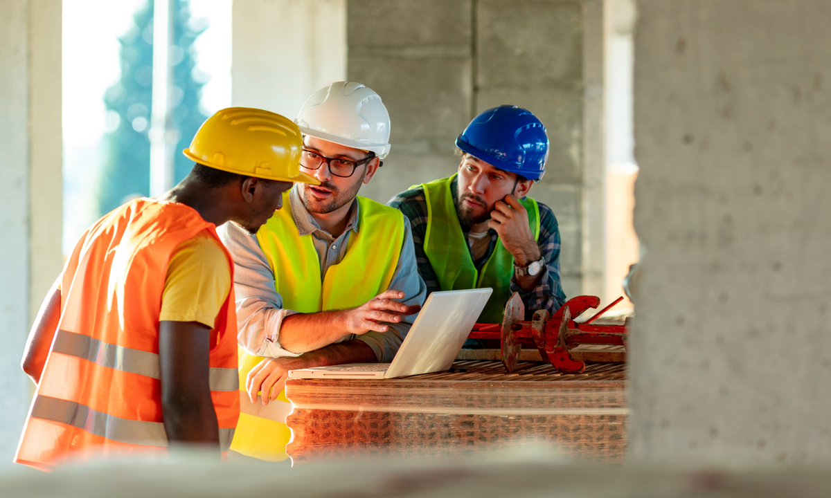 Three construction workers wearing safety helmets and high-visibility vests gather around a laptop on a workbench at an unfinished building site.