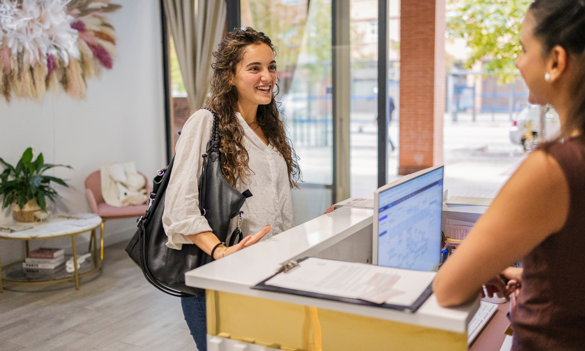 A woman in a white blouse and blue jeans stands at a reception desk, smiling and gesturing while talking to a receptionist inside a bright modern office or boutique.