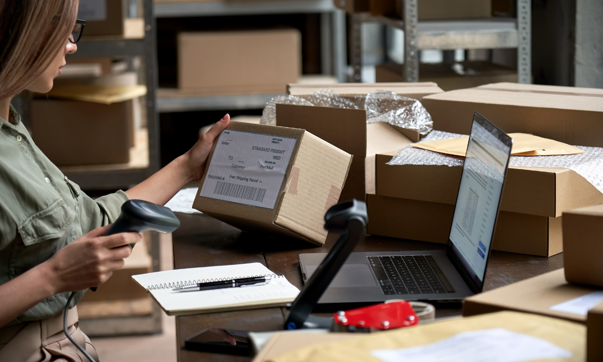 A woman in a warehouse scans a cardboard shipping box with a handheld barcode device while standing at a desk with a laptop, other packages, and shipping supplies.